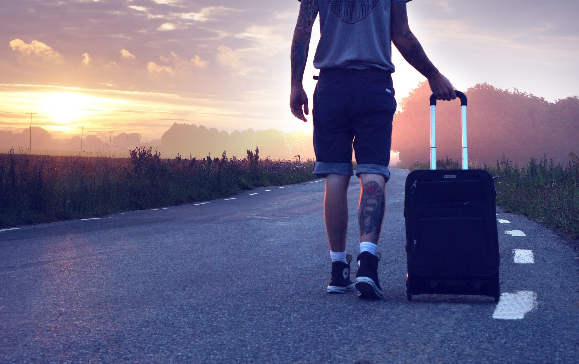 Chłopak z tatuażem na nodze ciągnie walizkę po drodze na tle zachodzącego słońca.